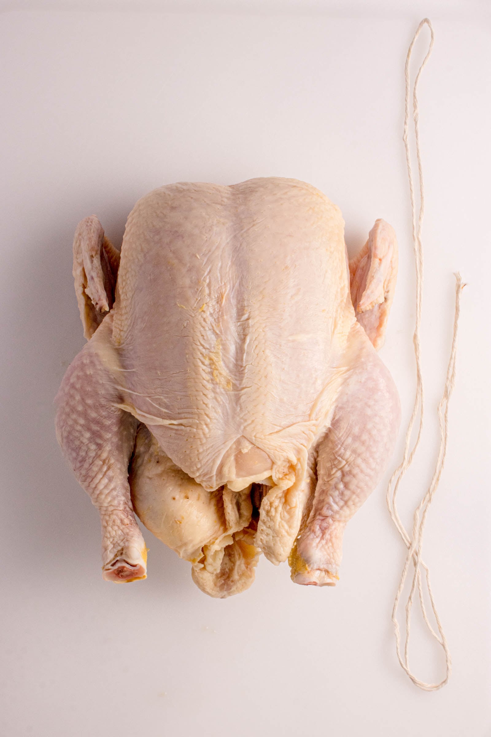 Raw whole chicken placed on a white surface next to a piece of kitchen twine.
