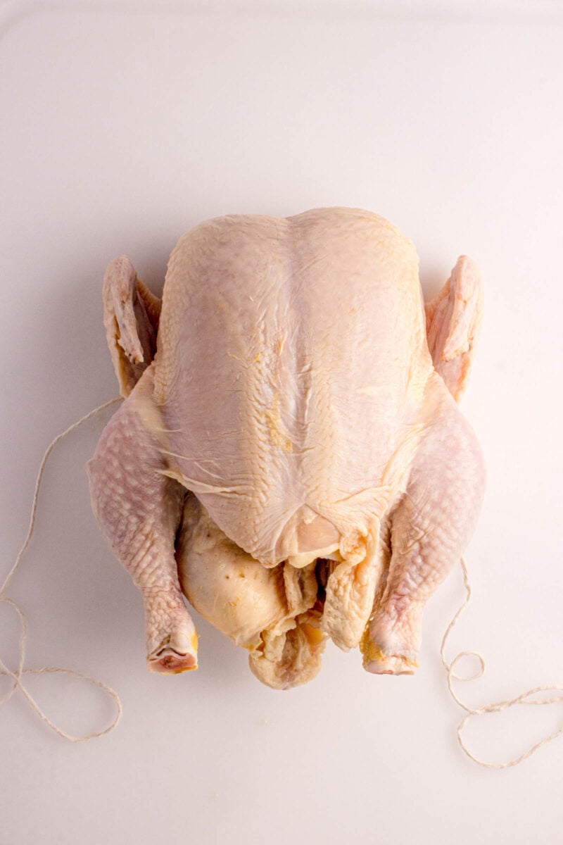 Raw whole chicken with tied legs placed on a white cutting board, viewed from above.