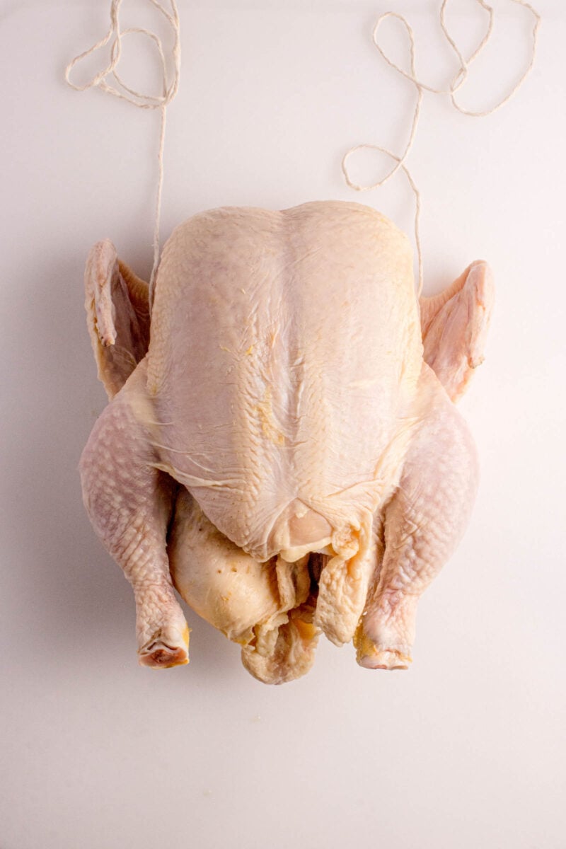 Raw whole chicken with wings and legs visible, lying breast-up on a white surface, with twine placed near the top.