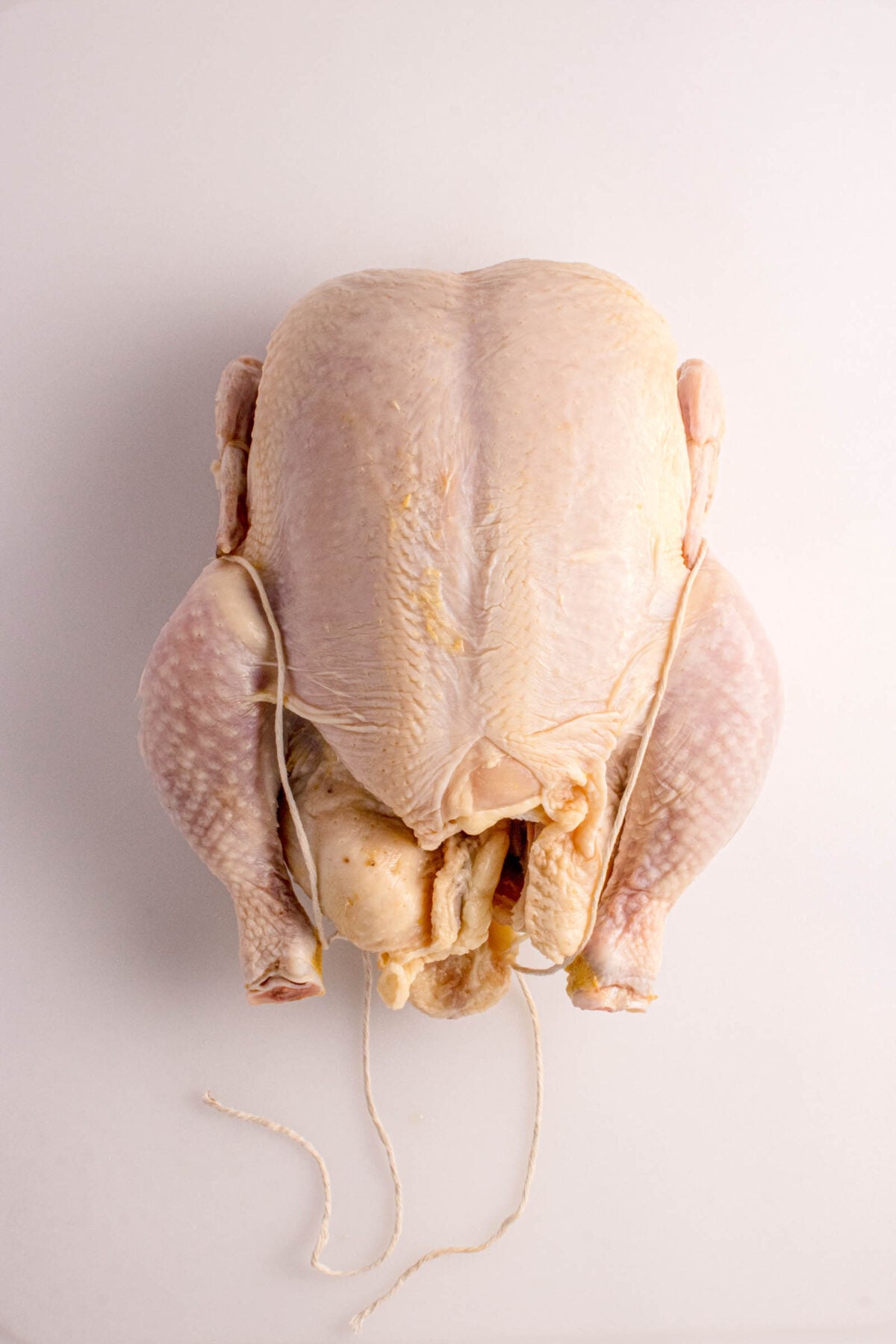 Raw whole chicken with legs tied together using string, placed on a plain white surface.