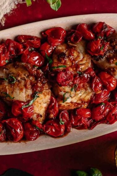Oval platter with cooked white fish fillets topped with roasted cherry tomatoes and herbs, placed on a red table.