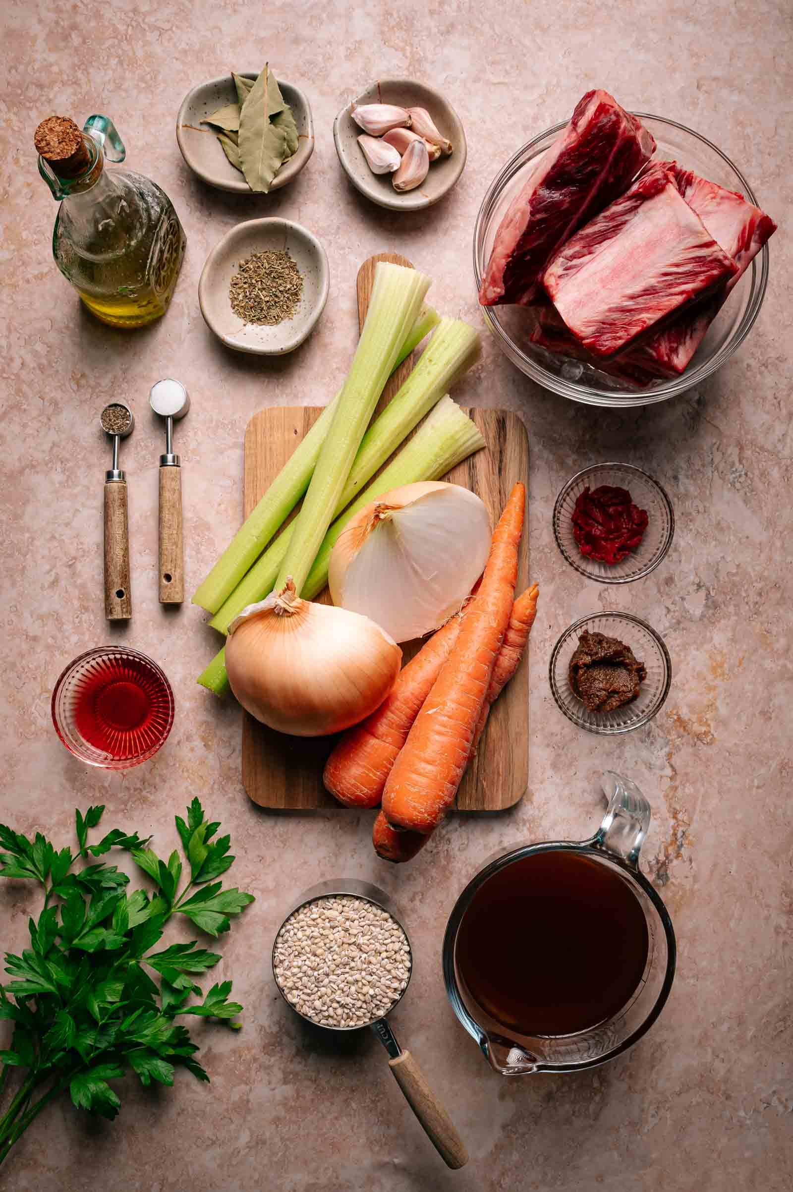 Various ingredients for a stew, including beef short ribs, carrots, celery, onions, garlic, parsley, spices, tomato paste, broth, barley, vinegar, and olive oil, arranged on a countertop.