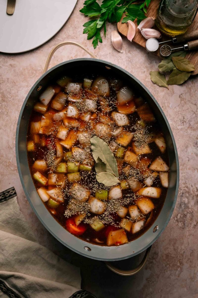 An oval pot filled with a stew mixture of chopped vegetables, broth, dried herbs, and bay leaves on a beige countertop with herbs and seasonings nearby.