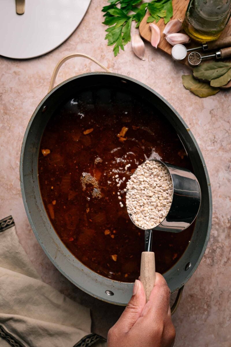 A hand pours a cup of uncooked barley into a pot of dark broth on a kitchen counter with herbs, garlic, oil, and bay leaves nearby.
