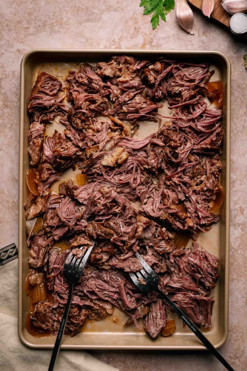 Shredded cooked beef spread on a baking sheet, with two serving forks resting in the meat.