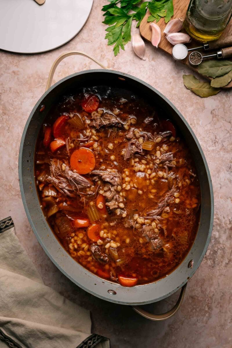 A pot of beef barley soup with chunks of beef, sliced carrots, celery, and barley in a rich broth sits on a countertop.