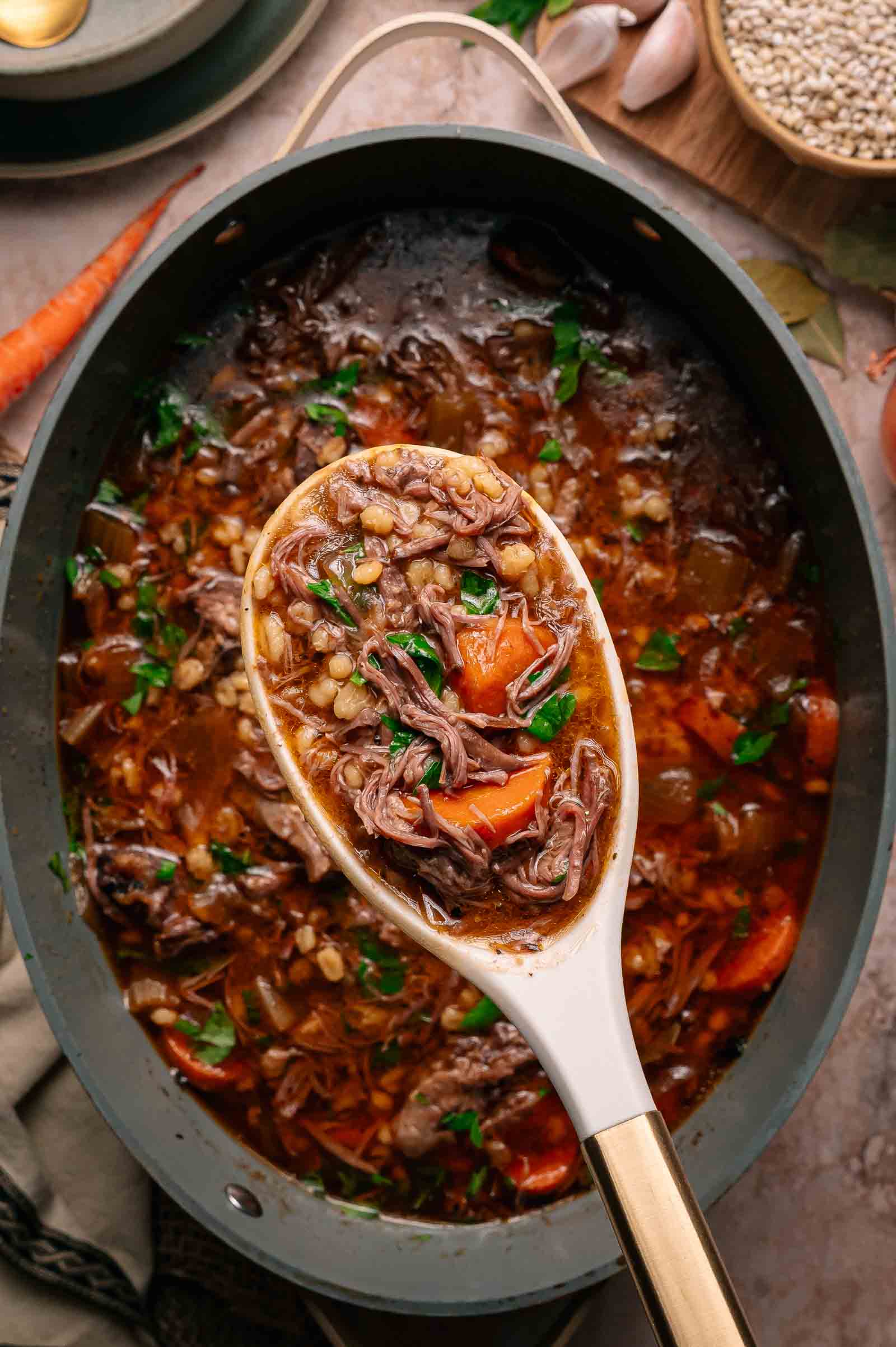 A spoonful of beef and barley stew with carrots and herbs is held above a pot filled with the same hearty stew.
