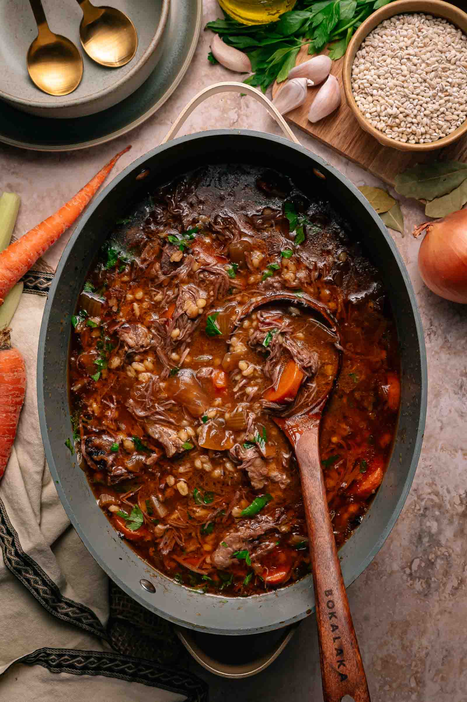 A pot of beef and barley soup with vegetables sits on a countertop; a wooden spoon rests in the pot. Surrounding ingredients include barley, garlic, carrot, and an onion.