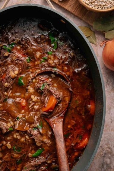 A pot of beef barley stew with chunks of meat, carrots, and barley, garnished with parsley, with a wooden spoon resting inside. An onion and bay leaves are nearby.