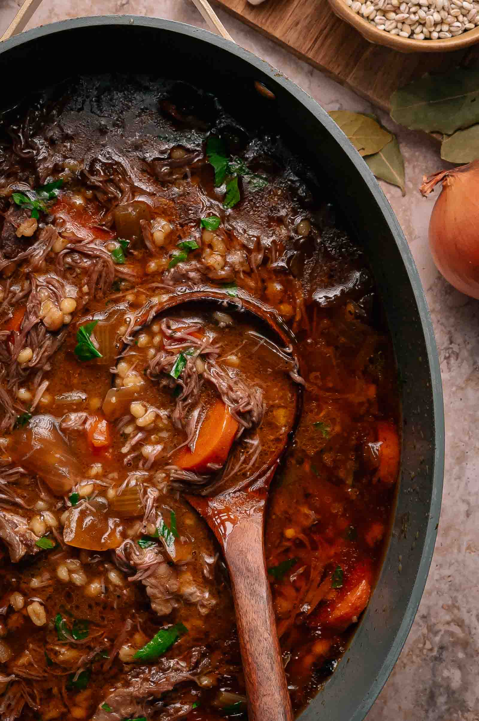 A pot of beef barley stew with chunks of meat, carrots, and barley, garnished with parsley, with a wooden spoon resting inside. An onion and bay leaves are nearby.