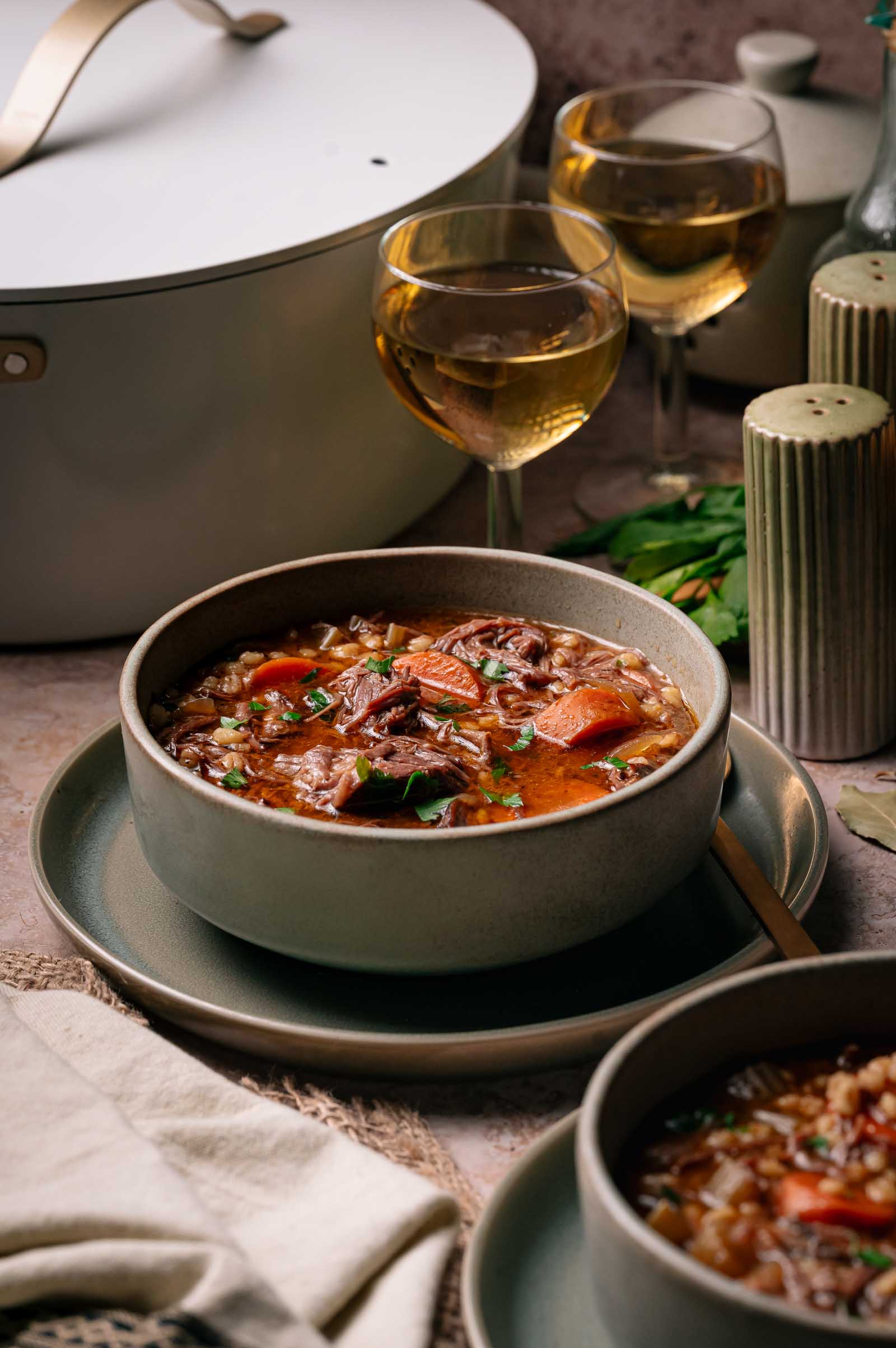 A bowl of beef stew with vegetables is served on a plate with a spoon, accompanied by two glasses of white wine and a pot in the background.