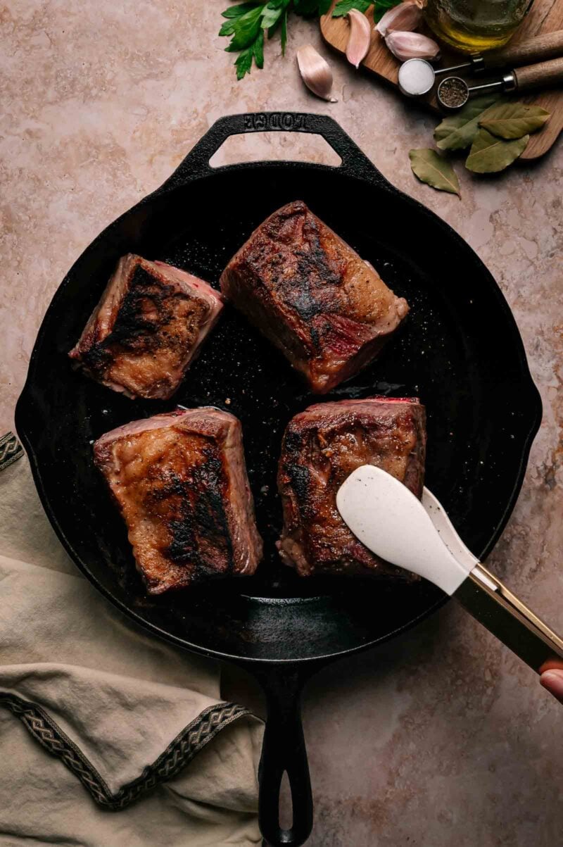 Four browned pieces of meat are searing in a black cast iron skillet, with a spatula flipping one piece; ingredients and kitchen towel are nearby on a beige countertop.
