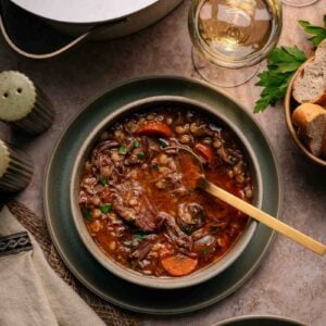 A bowl of beef and vegetable soup with a spoon, surrounded by bread, wine glasses, salt and pepper shakers, and fresh parsley on a table.