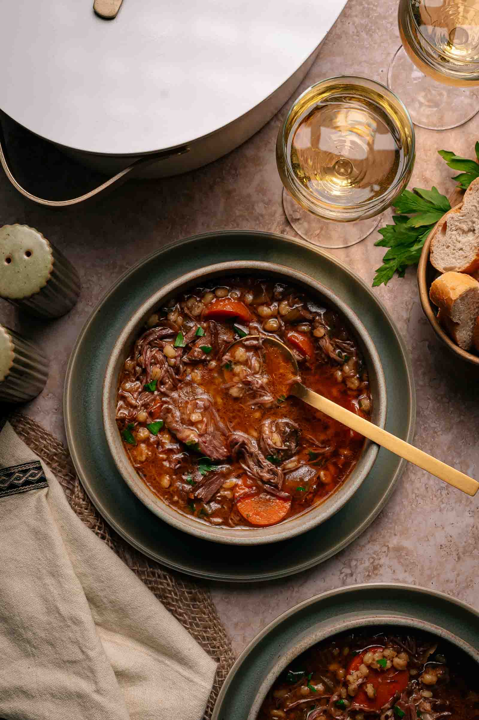A bowl of beef and vegetable soup with a spoon, surrounded by bread, wine glasses, salt and pepper shakers, and fresh parsley on a table.