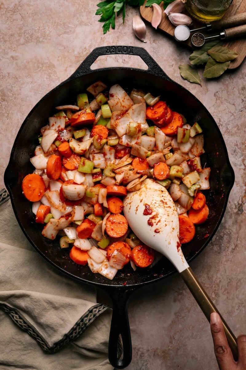 A hand stirs chopped carrots, celery, and onions in a black cast iron skillet with a white spatula.