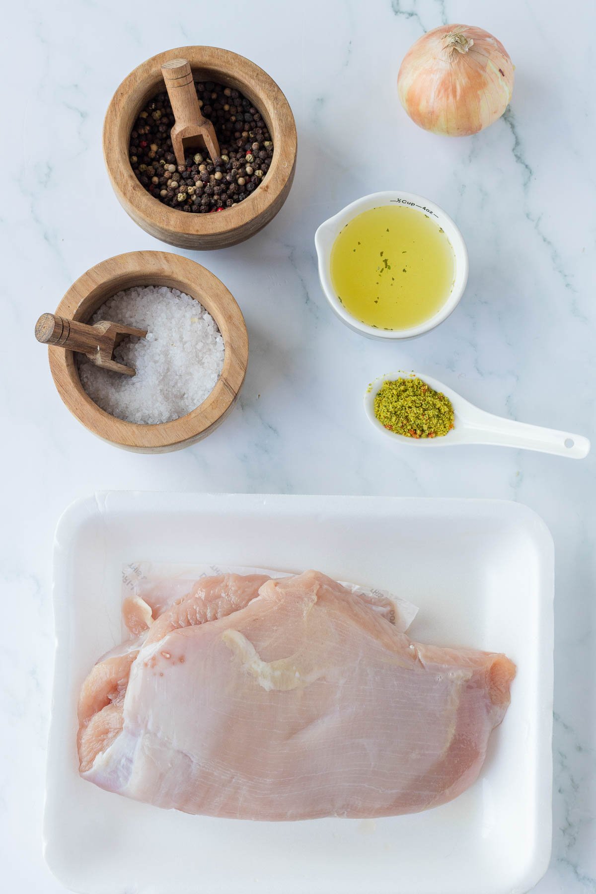 Raw turkey breast on a white tray, with bowls of salt, peppercorns, a small pitcher of oil, a yellow spice blend on a spoon, and an onion on a marble surface.