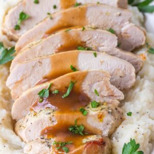 Sliced turkey breast with brown gravy served over mashed potatoes, garnished with chopped parsley on a white plate.