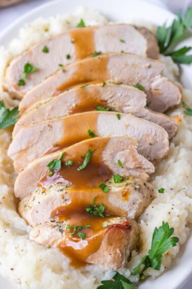 Sliced turkey breast with brown gravy served over mashed potatoes, garnished with chopped parsley on a white plate.