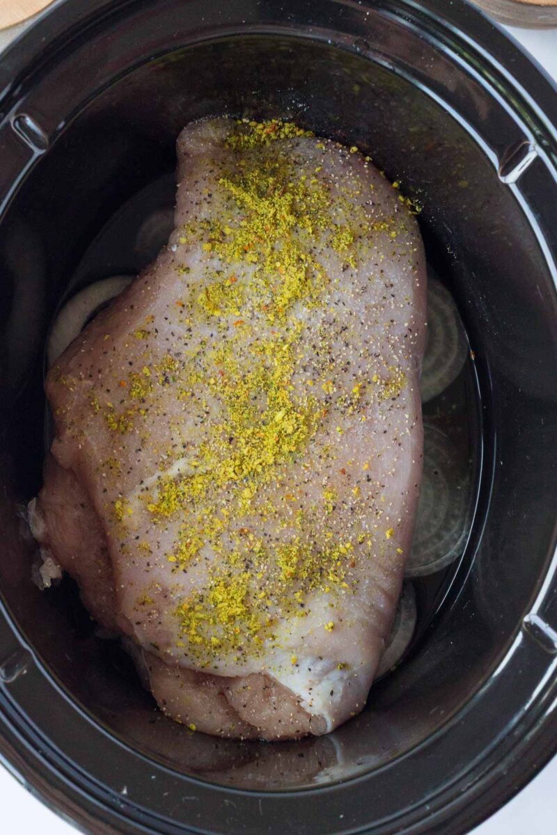 Raw seasoned turkey breast placed inside a slow cooker, with visible sliced onions underneath and some liquid at the bottom.
