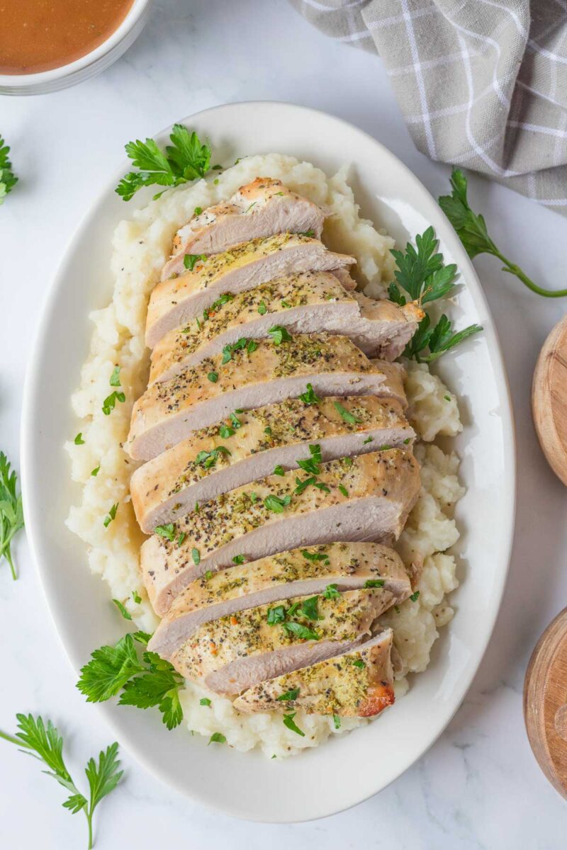 Sliced boneless turkey breast served on a bed of mashed potatoes, garnished with chopped parsley on a white oval plate.
