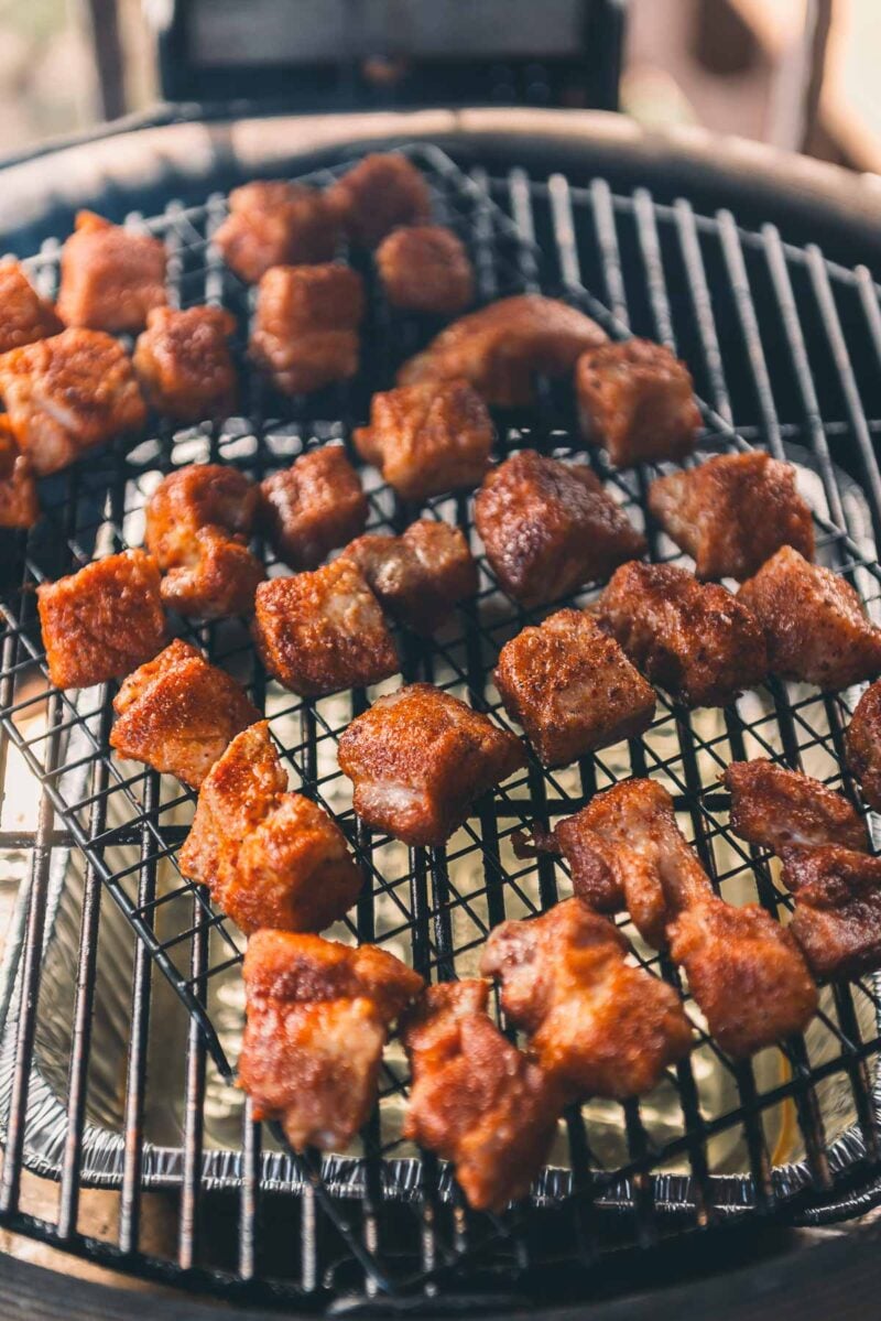 Cubed pieces of seasoned pork belly are smoking on a barbecue grate.