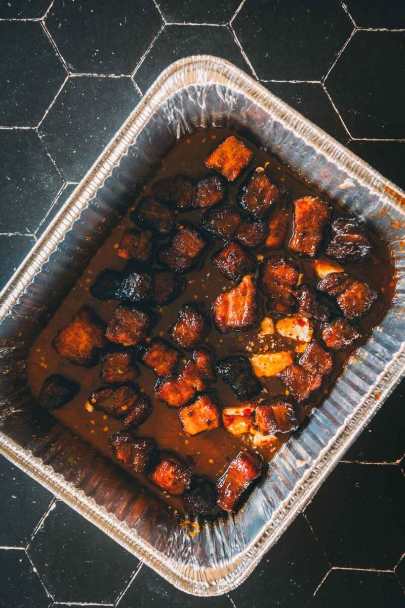 Aluminum tray containing glazed, cubed pieces of smoked pork belly burnt ends in sauce, placed on a black tile surface.