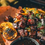 Glazed smoked pork belly burnt ends garnished with microgreens, served with charred orange slices and a side of maple chipotle bourbon sauce on brown parchment paper.