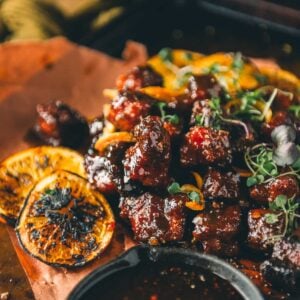 Glazed smoked pork belly burnt ends garnished with microgreens, served with charred orange slices and a side of maple chipotle bourbon sauce on brown parchment paper.