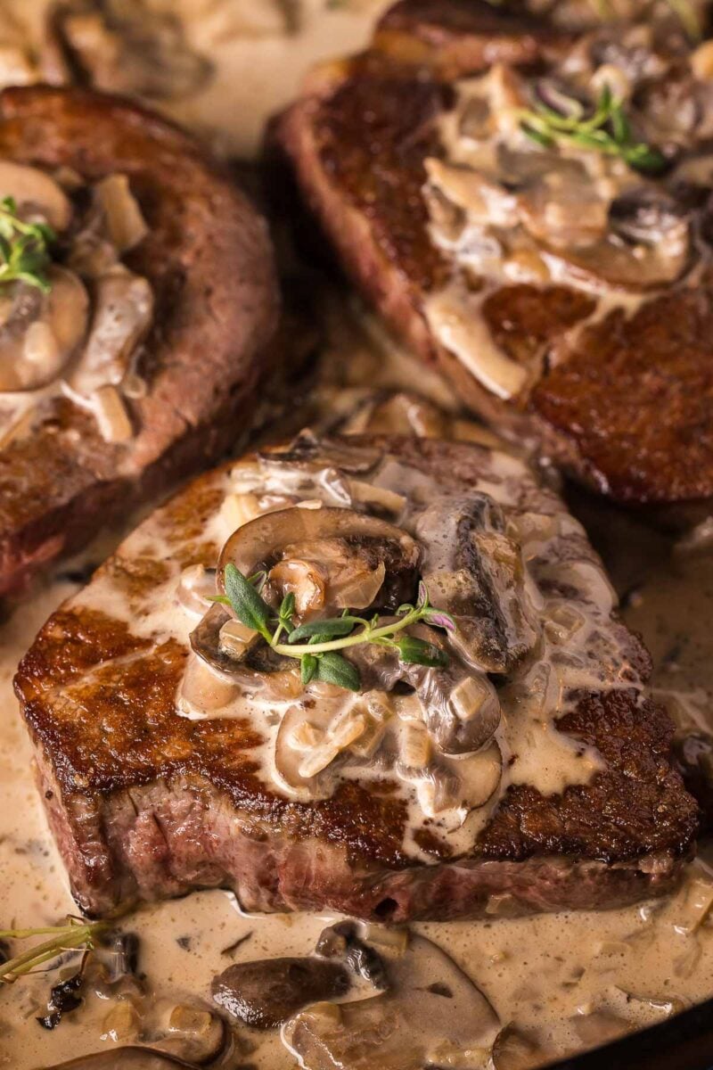 Close-up of seared steak topped with creamy mushroom sauce and garnished with fresh herbs.