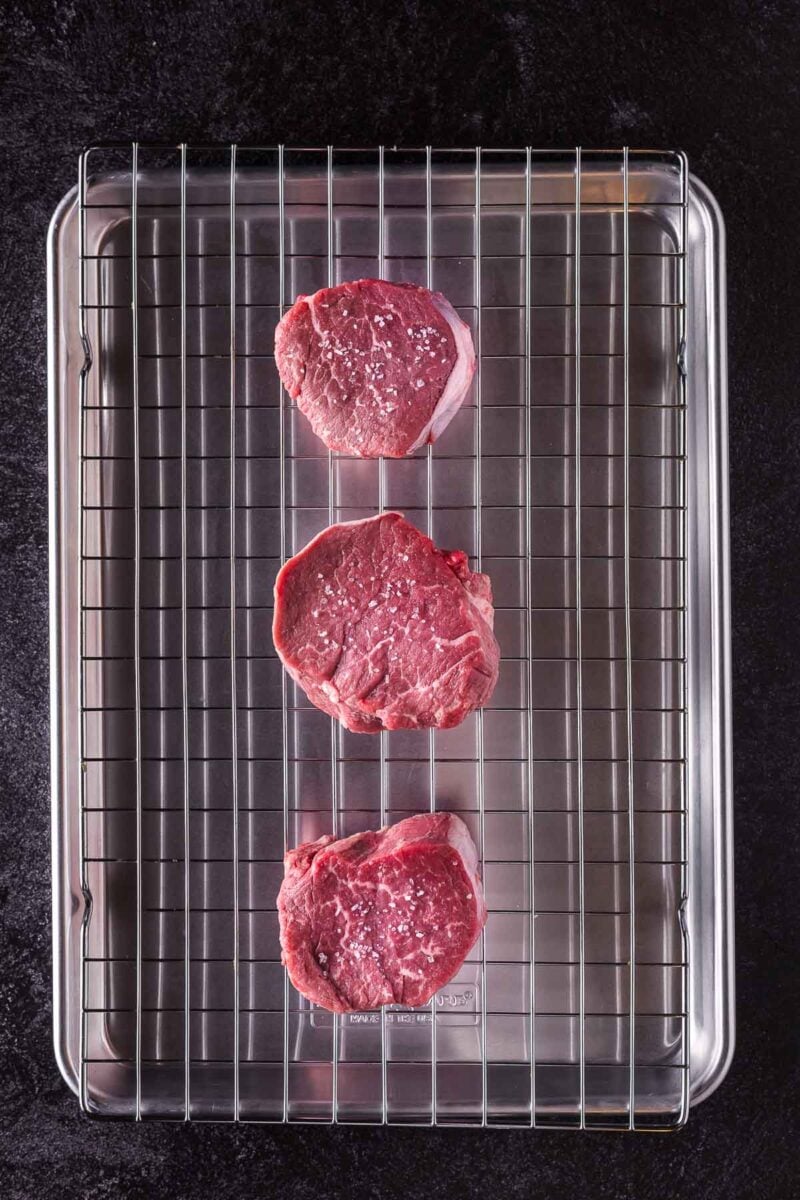 Three raw filet mignon steaks sprinkled with salt are arranged on a wire rack over a baking sheet on a dark surface.