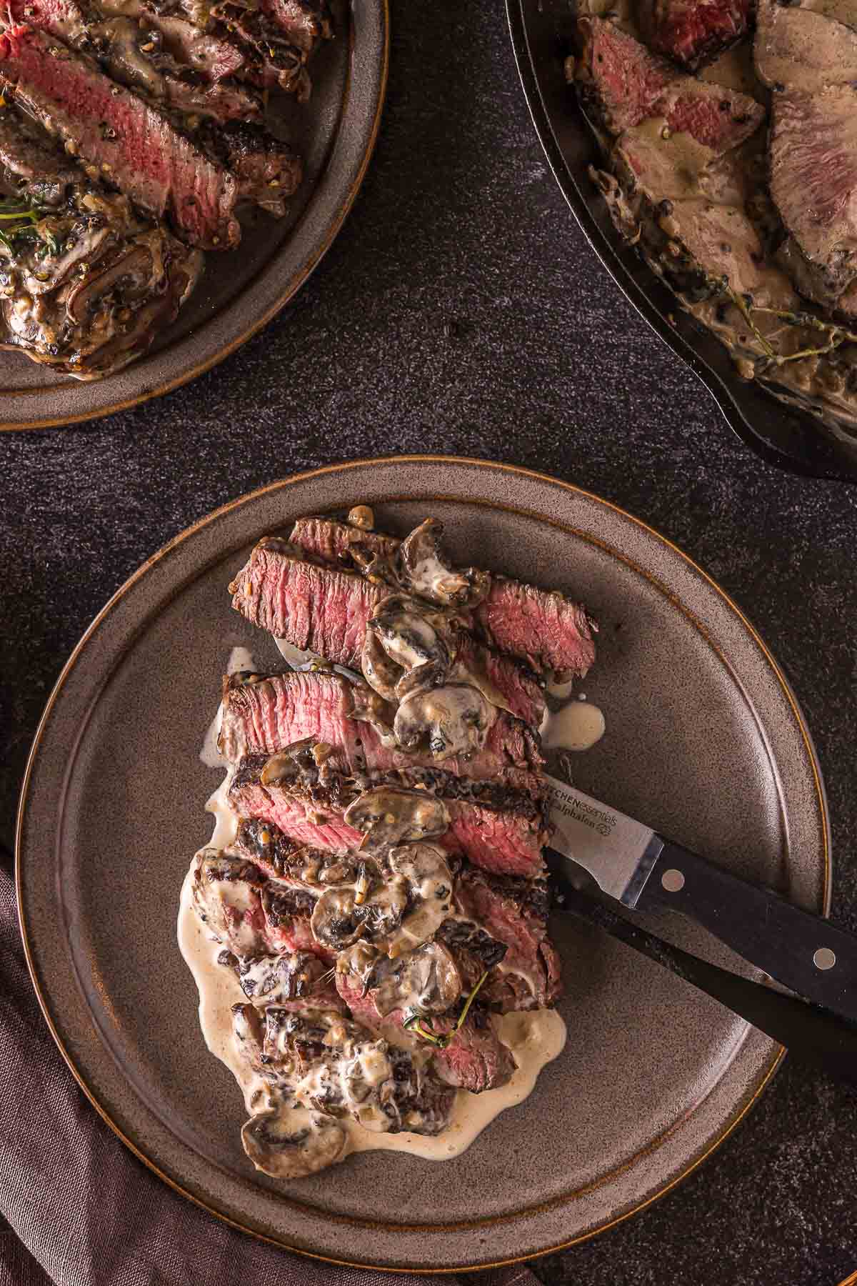 Sliced steak topped with creamy mushroom sauce on a ceramic plate, with a knife resting on the side, and more steak visible in the background.