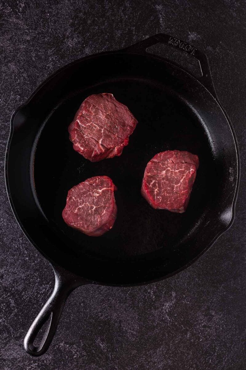 Three raw filet mignon steaks are placed in a black cast iron skillet on a dark textured surface.