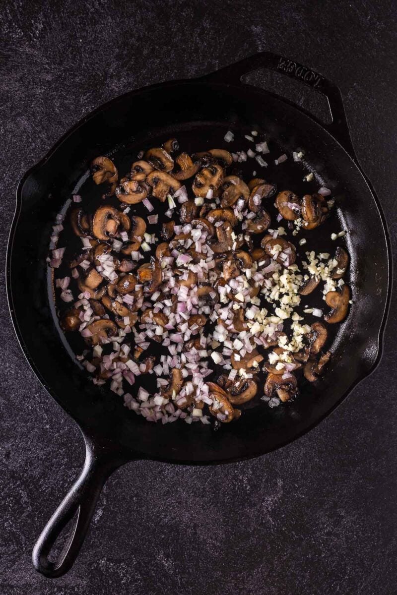 Sliced mushrooms, chopped shallots, and minced garlic cooking in a black cast iron skillet on a dark surface.