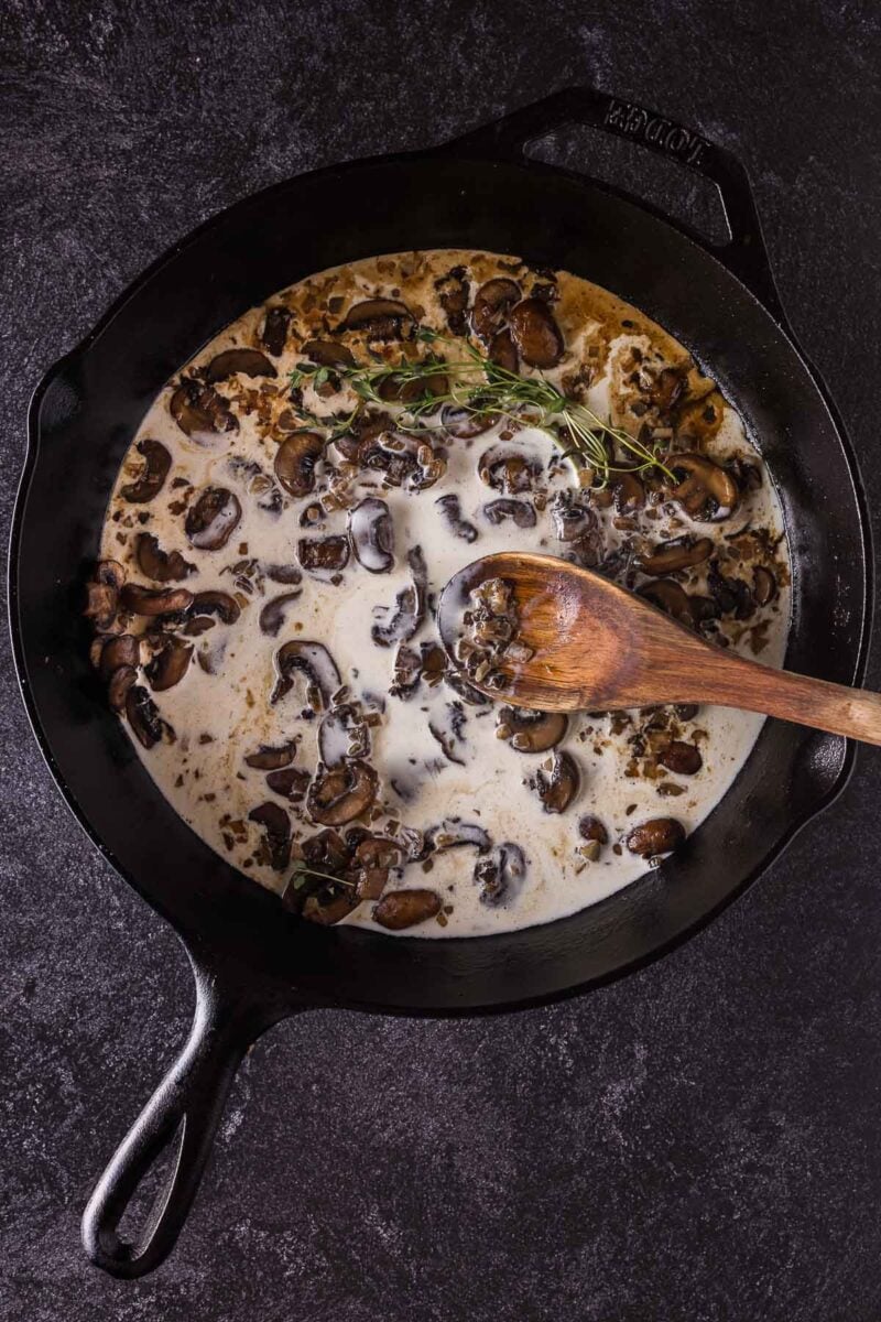 A cast iron skillet with creamy mushroom sauce, garnished with fresh thyme, being stirred with a wooden spoon on a dark surface.