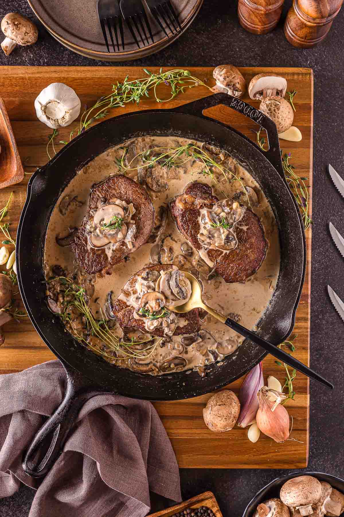 Cast iron skillet with seared steak in creamy mushroom sauce, garnished with herbs, surrounded by raw mushrooms, shallots, and utensils on a wooden board.