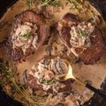 Three seared steaks in a skillet topped with creamy mushroom sauce and garnished with fresh thyme, with a spoon resting on one steak.