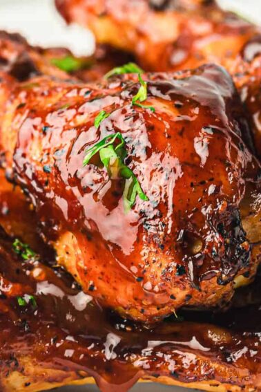 Close-up of grilled chicken drumsticks coated in barbecue sauce and garnished with chopped herbs, served on a white plate.