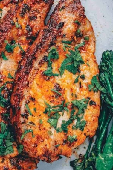 Three seasoned, cooked pork chops garnished with chopped herbs are served next to a portion of steamed broccolini on a plate.