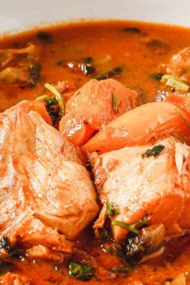 A bowl of fish stew with chunks of fish, tomatoes, herbs, and vegetables in a reddish broth.