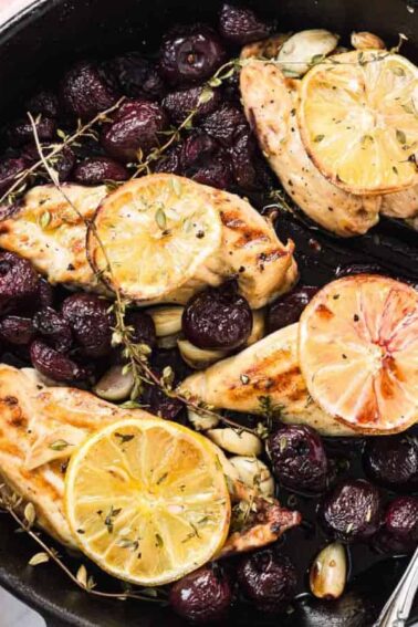 Cast iron skillet with roasted chicken breasts topped with lemon slices, surrounded by roasted grapes, garlic cloves, and sprigs of thyme. A serving spoon rests in the skillet.