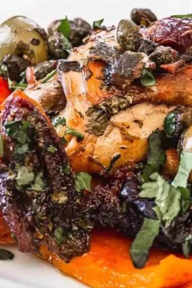 Roasted chicken with olives, capers, herbs, and prunes served over roasted sweet potato wedges on a white plate with a fork.