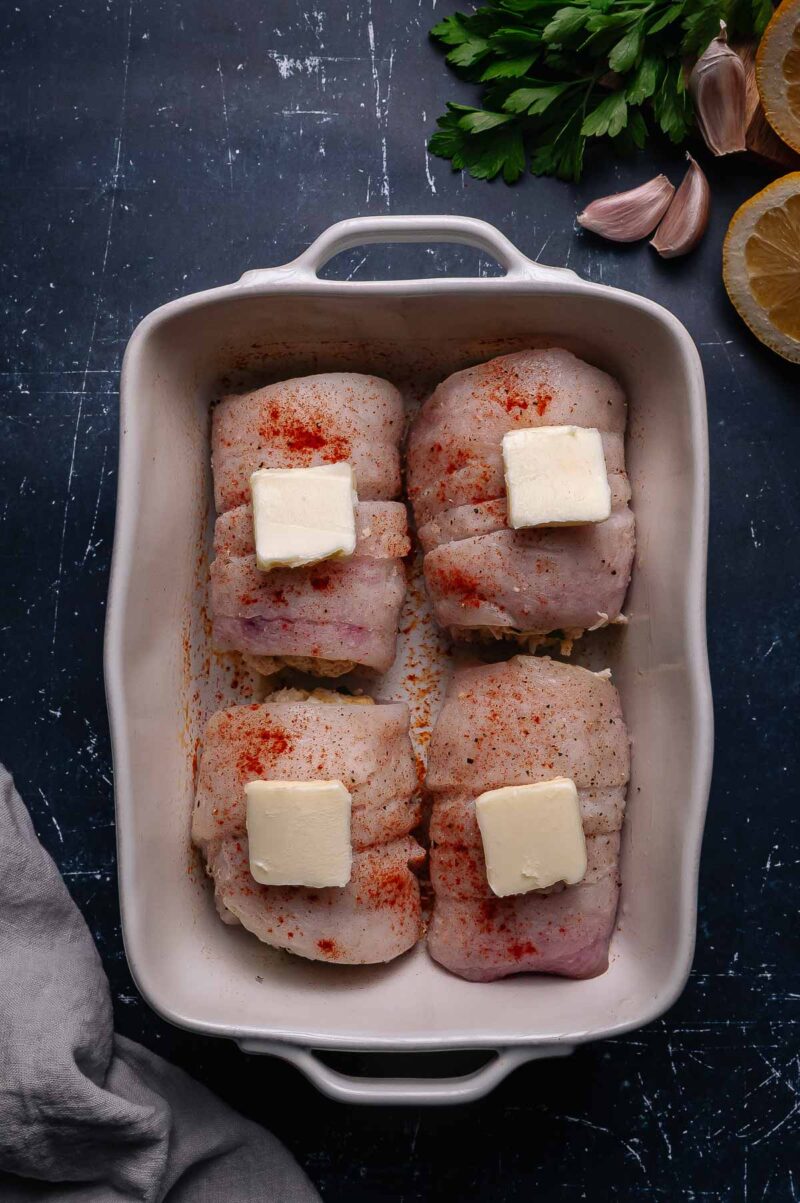 Four pieces of raw seasoned white fish fillets in a baking dish, each topped with a pat of butter. Fresh parsley, garlic, and lemon are on the side.