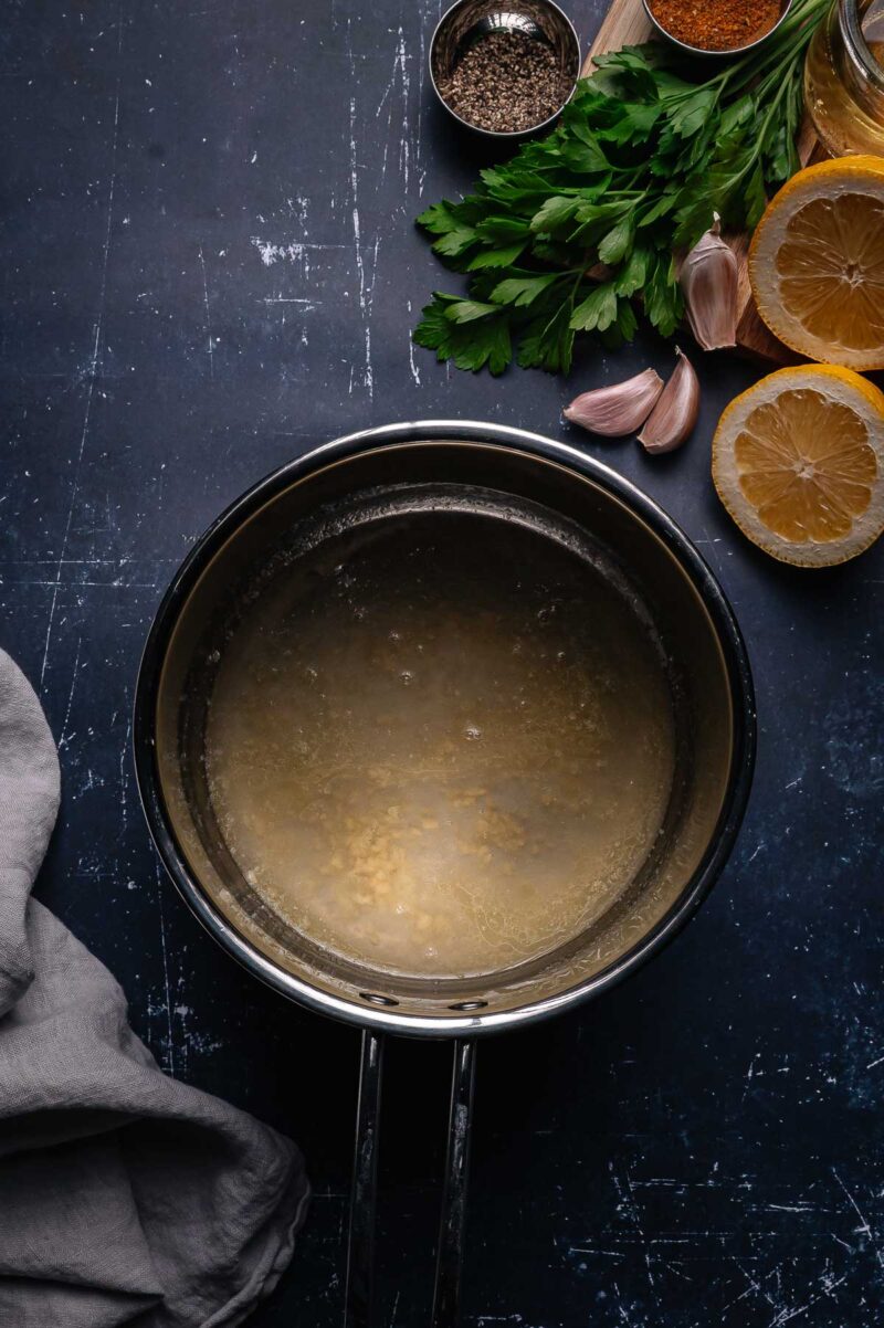 A saucepan with melted butter sits on a dark counter next to lemon slices, garlic cloves, fresh herbs, spices, and a gray cloth.
