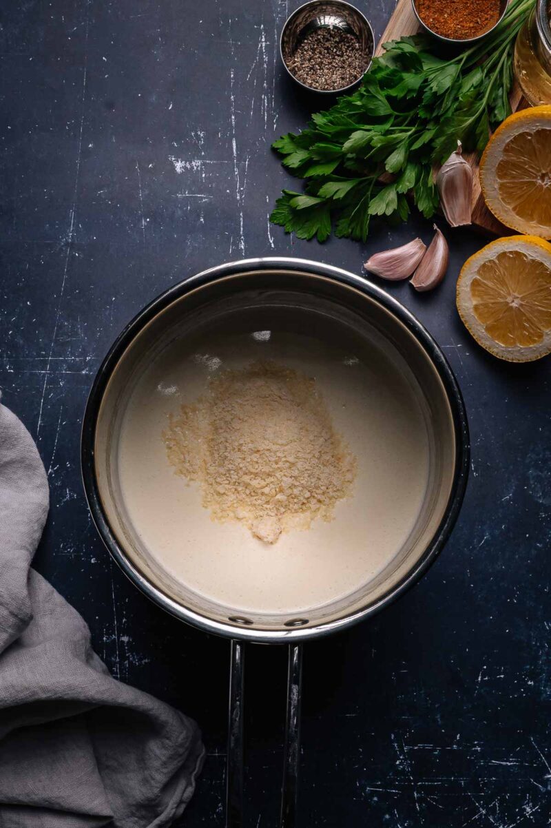 A saucepan with cream and grated cheese, next to parsley, garlic cloves, lemon halves, and small bowls of spices on a dark surface.