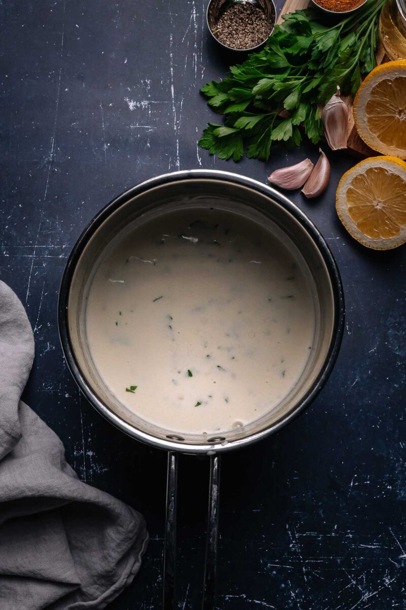 A saucepan filled with creamy white wine sauce, surrounded by fresh parsley, garlic cloves, lemon slices, black pepper, and a gray napkin on a dark surface.