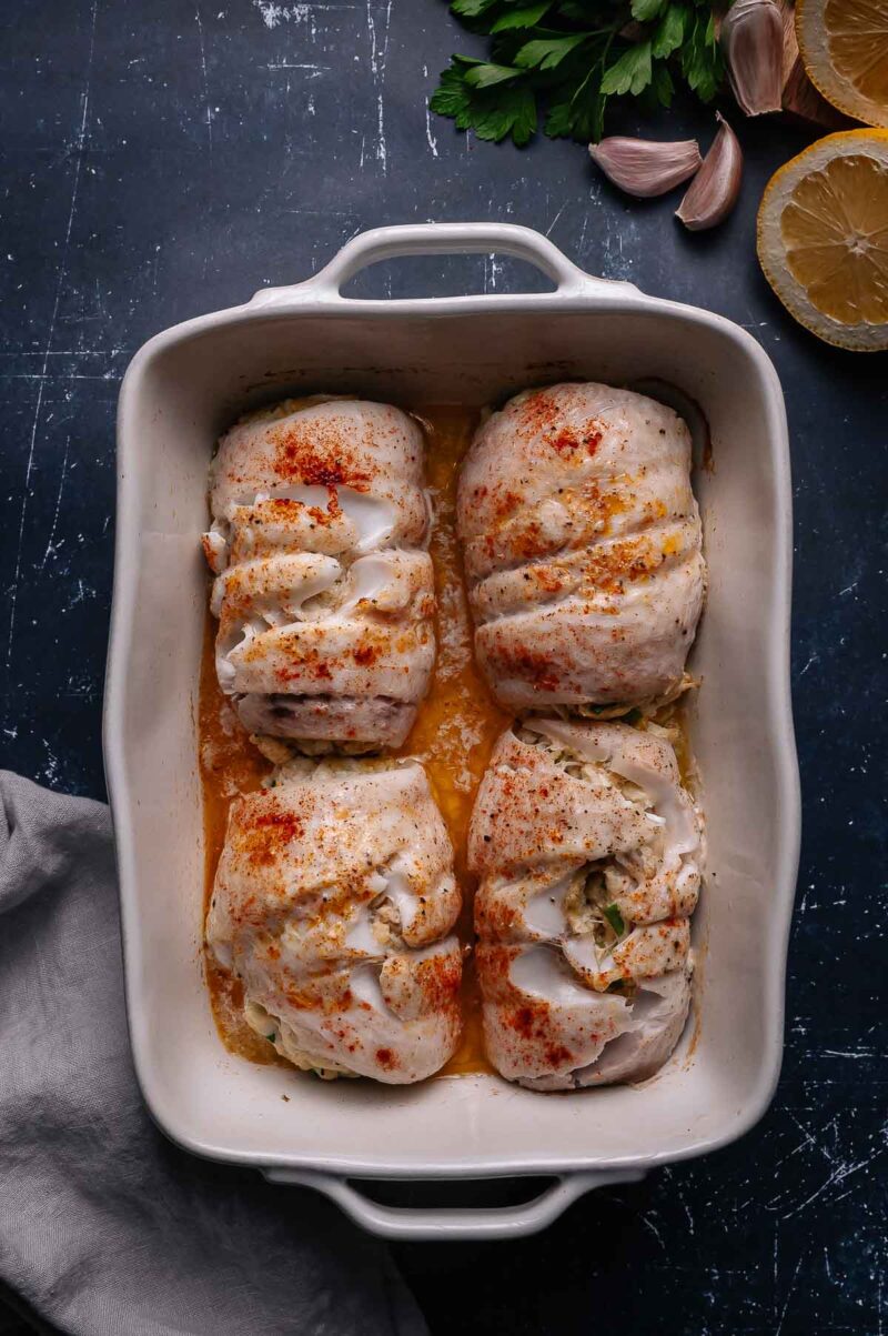 Four baked stuffed flounder filets in a white ceramic baking dish, surrounded by garlic, lemon halves, and parsley on a dark surface.