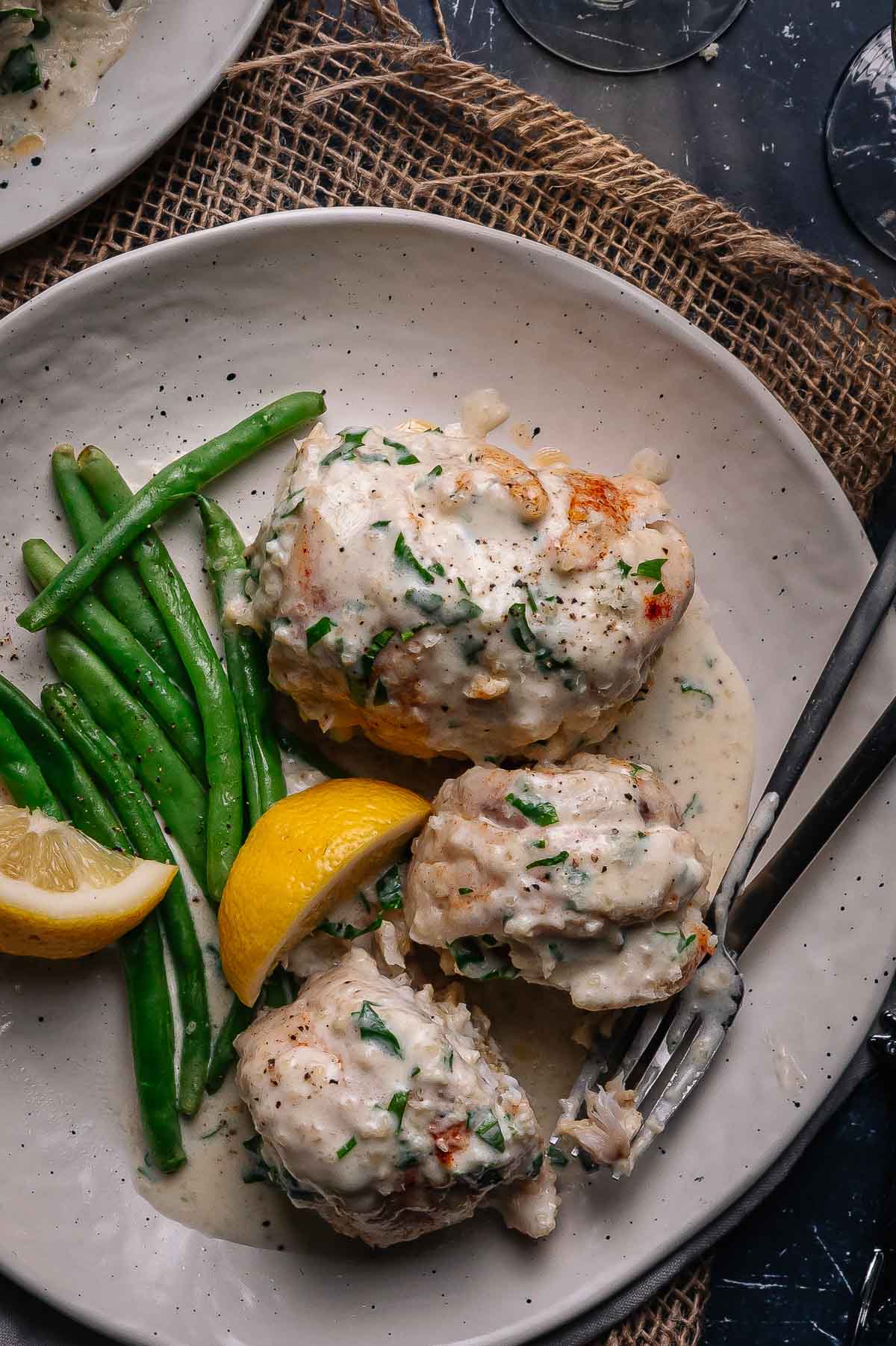 Plate of creamy baked crab stuffed flounder with herbs, served with green beans and lemon wedges on a white dish, with a fork and knife on the side.