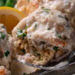 A close-up of a creamy baked crab stuffed flounder garnished with chopped herbs, served with a lemon wedge on a white plate.