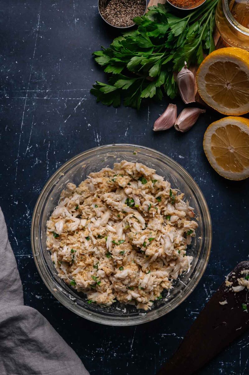 A glass bowl filled with prepared crab meat stuffing sits on a dark surface, surrounded by parsley, garlic cloves, lemon halves, seasonings, and a gray cloth.