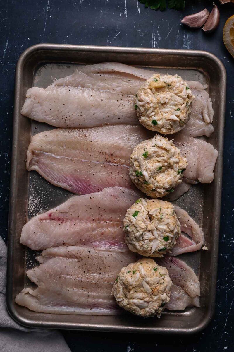 A metal tray with four raw flounder fillets and four uncooked crab stuffing arranged side by side.